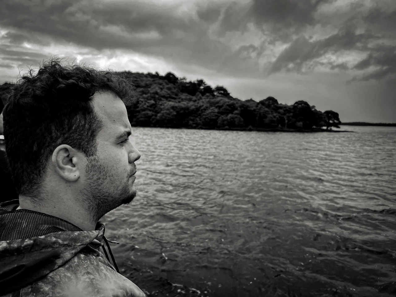 Black-and-white side profile of a person beside a lake, with a wooded shore behind.