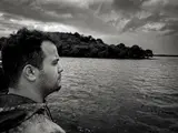 Black-and-white side profile of a person beside a lake, with a wooded shore behind.