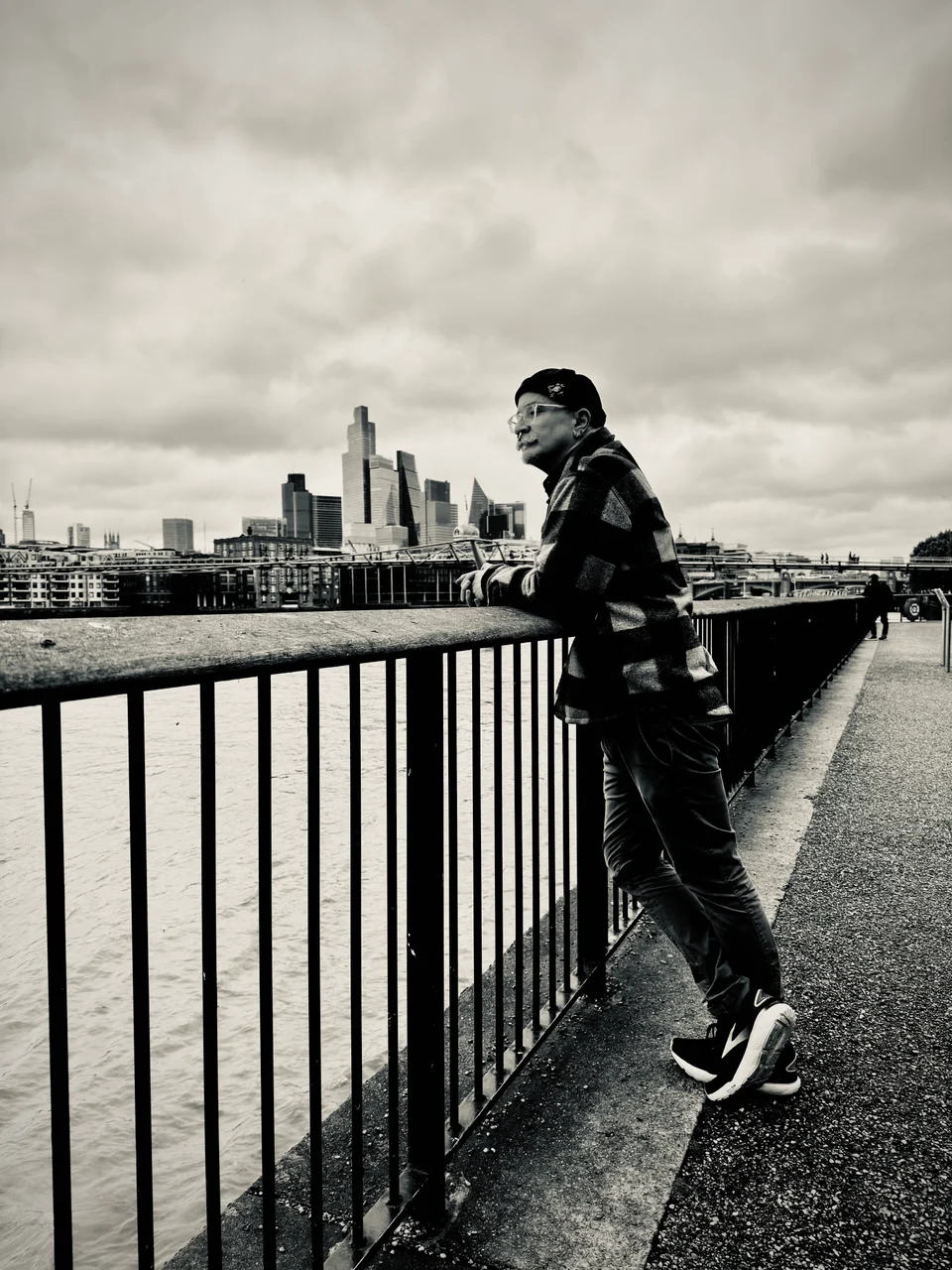 Black-and-white photograph of a person leaning against a riverside railing, with a city skyline across the water.