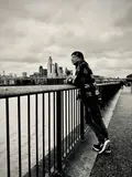 Black-and-white photograph of a person leaning against a riverside railing, with a city skyline across the water.