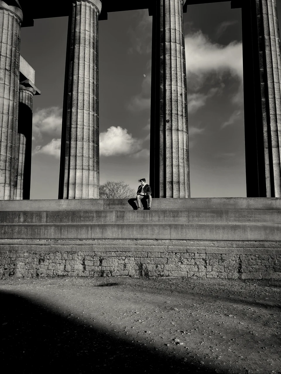 Black-and-white photograph of a small figure seated on steps between tall classical columns.