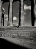 Black-and-white photograph of a small figure seated on steps between tall classical columns.