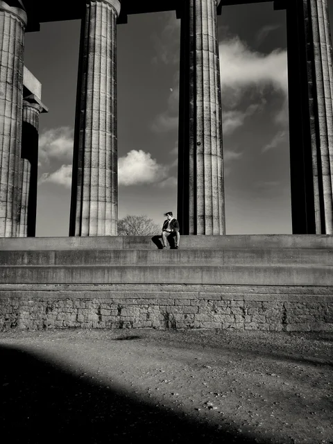 Black-and-white photograph of a small figure seated on steps between tall classical columns.