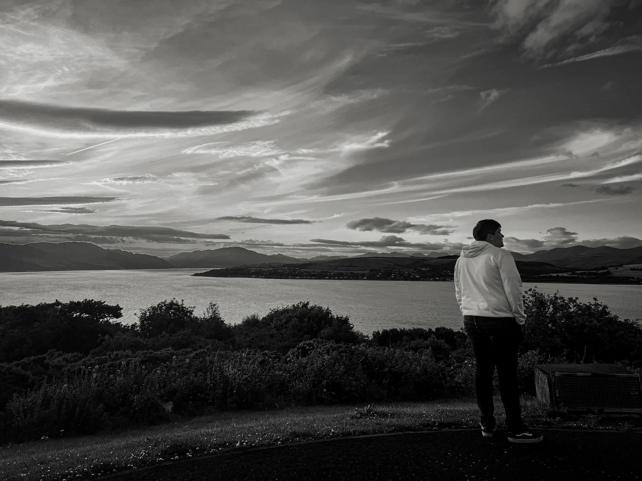 Black-and-white photograph of a person facing a lake and distant hills beneath streaked clouds.