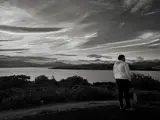 Black-and-white photograph of a person facing a lake and distant hills beneath streaked clouds.